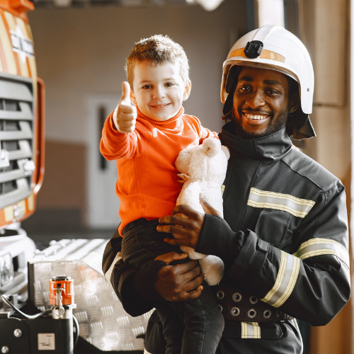 Firefighter holding a young boy who is giving a thumbs up hand gesture